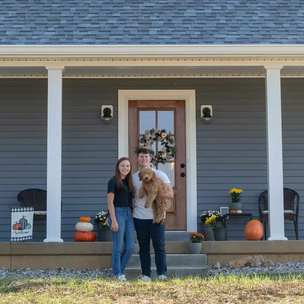 Pumpkin and fall decorations on porch with two people and dog.