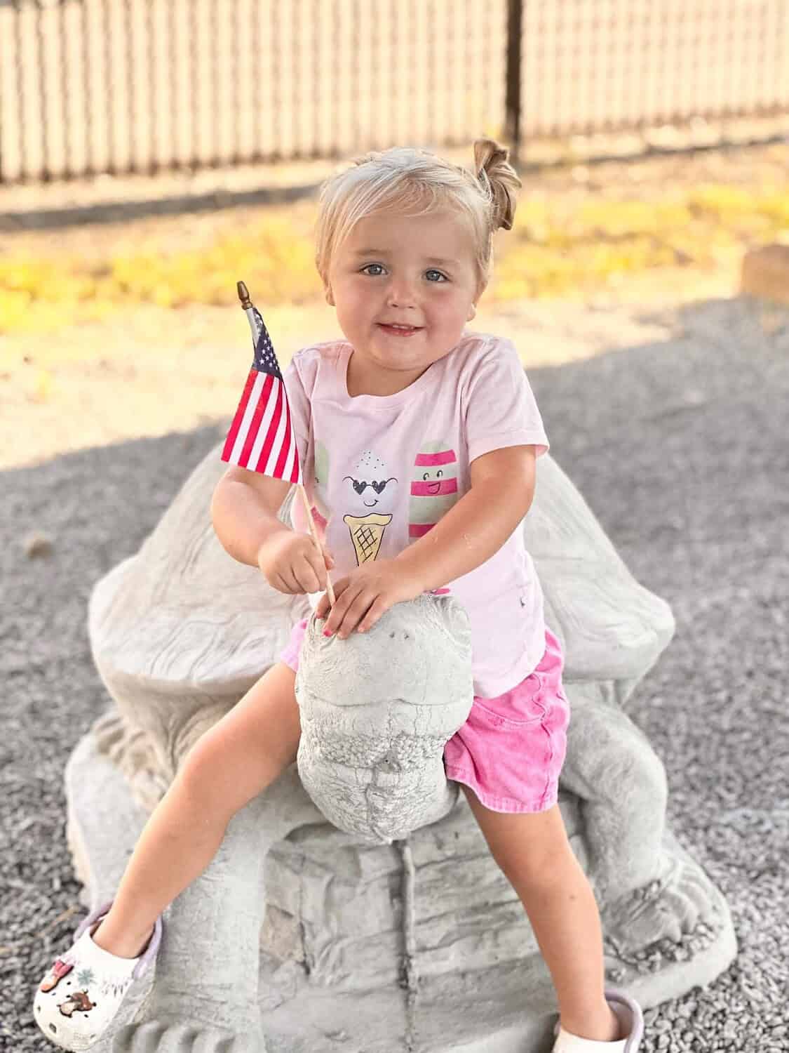 Young girl with blonde hair, pink shorts, and a white t-shirt sitting on a stone lion statue.
