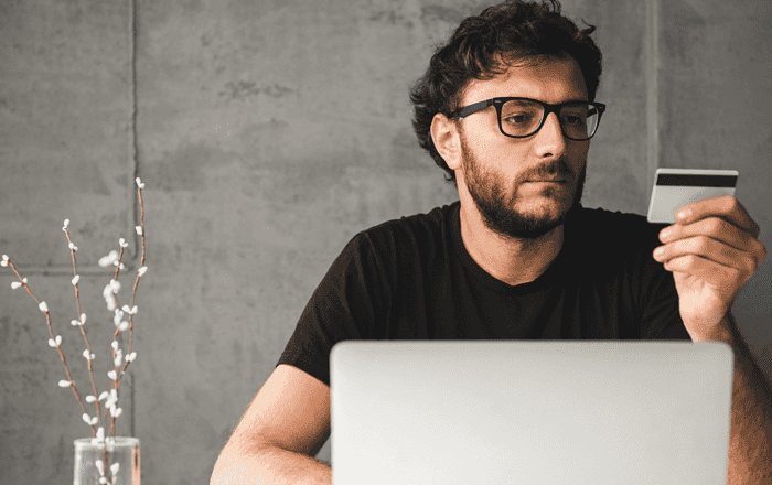 Man looking at smartphone with laptop in front and plant on side.