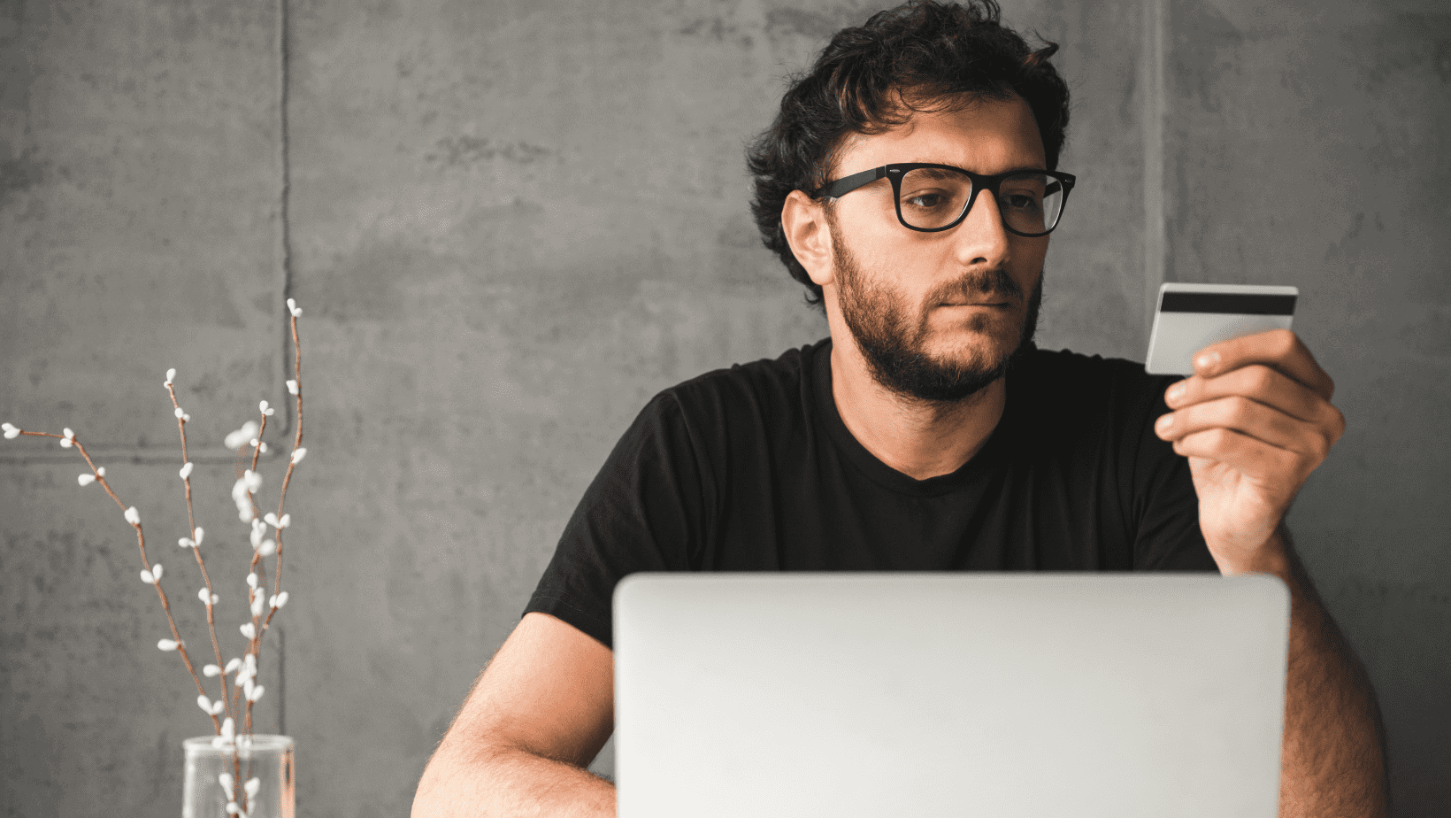 Man looking at smartphone with laptop in front and plant on side.