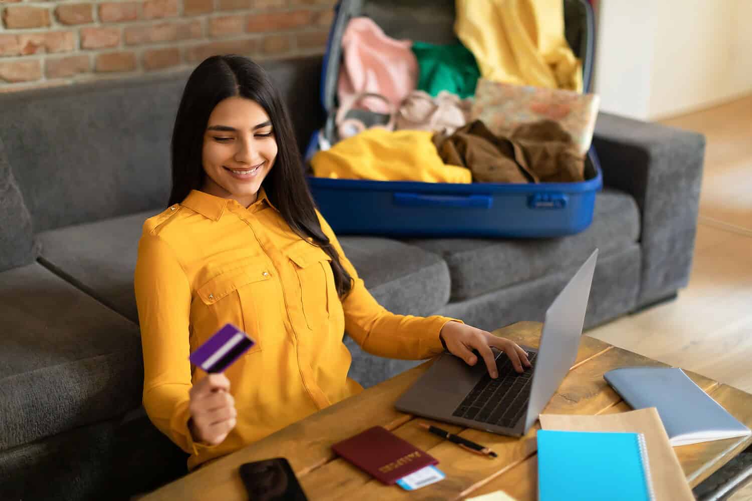 Positive,Spanish,Lady,Using,Credit,Card,And,Laptop,,Booking,Abroad Woman packing clothes with credit card and laptop at home.