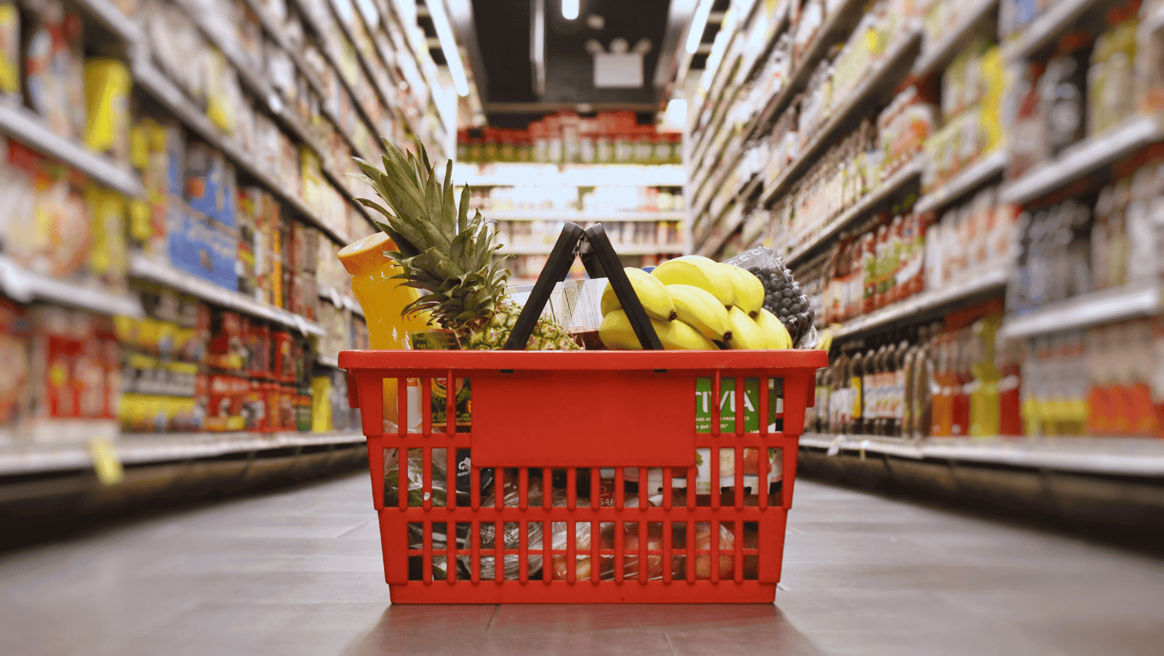 Pineapple, bananas, and assorted fruits in a red shopping basket on supermarket aisle floor.